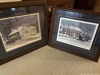 Photo of two framed art prints leaning against wall, winter scenes with snow, trees, and historic houses.