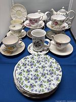 Porcelain tea set with various floral-designed teacups, saucers, two teapots, a mug, and four floral-patterned plates on a blue fabric background.