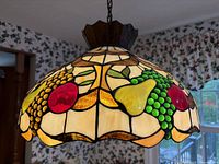 Close-up view of the stained glass pendant light showing colorful fruit patterns with grapes, pears, and apples in red, green, yellow, and cream colors. The scalloped edges and leaded glass detail are visible.