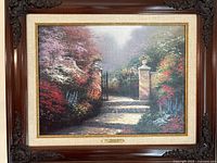 Framed Thomas Kinkade canvas painting depicting a garden gate with blooming trees and shrubs, soft evening lighting, dark wood frame with ornamental corners.