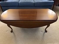 View of traditional drop-leaf wood coffee table with functional fold-down leaf, brown polished surface, placed on carpet in front of sofa.