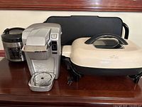 Photo showing four kitchen appliances: silver coffee maker, black digital rice cooker, cream Rival electric skillet with lid, and black electric griddle behind them.