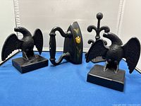 Two cast iron eagle bookends, a black sad iron with floral decoration, and a black decorative stand on a blue cloth.