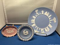 Three Wedgwood ceramic plates displayed against neutral background. Large blue plate with white relief figures, medium blue plate with wreath design, small pink plate with relief decoration.