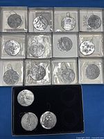 Photo shows all 15 Wendell August metal Christmas ornaments, arranged in clear plastic boxes with black backgrounds. The ornaments have different embossed holiday patterns including Santa, stockings, holly leaves, nativity scene, candles, angels, and snowflakes. Three additional ornaments are outside their boxes at the bottom.