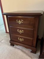Full view of wooden nightstand showing three drawers with ornate brass handles, traditional carved feet, and polished wood surface.