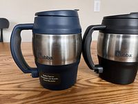 Blue and black Bubba insulated travel mugs side by side on wooden table showing stainless steel bodies and handles with bottle openers.