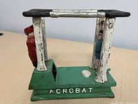 Side view of acrobat piggy bank showing gymnast figurine hanging from silver horizontal bars, green base with text ACROBAT in white, and figure in red.