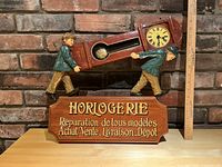 Full front view of the wooden clock sign on a shelf with a brick background, showing two figures carrying the clock case and the French text base. A ruler shows the height approximately 16 inches.