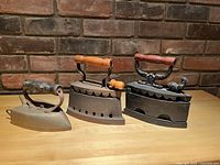 Set of three antique cast iron sad irons with visible wooden handles on a wooden surface against a brick background