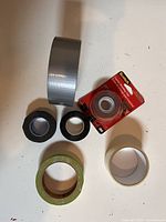 Photo showing several rolls of tape including a large roll of silver duct tape, two rolls of black electrical tape, a roll of clear Scotch tape in packaging, and a roll of green masking tape.