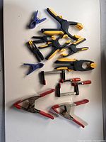 Photo showing assortment of clamps including black and yellow plastic spring clamps, blue plastic clamps, red-handled locking pliers, and red-handled bar clamps on a white surface.