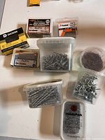 Photo shows several plastic tubs and boxes containing assorted nails and screws of various sizes and types laid out on a white surface.