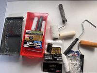 Photo showing black metal paint tray, two red plastic paint trays, paint rollers, unopened foam roller covers and paint can opener.