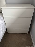 Front view of the white 4-drawer dresser next to a bed, showing clean modern design and integrated handles