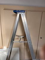 Full view of the aluminum 6-foot A-frame ladder showing silver legs and blue plastic top tray against beige wall.