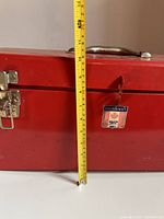 Front view of red Mastercraft toolbox with measuring tape showing height approximately 10 inches. Shows metal handle and Mastercraft badge with slight rust and wear.