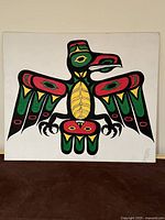 Frontal photo of the original Thunderbird painting displayed on a brown surface against a light wall. The painting shows a bold, colorful indigenous Thunderbird design on white canvas.