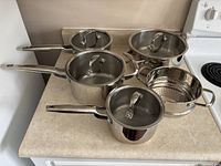 Five stainless steel pots and a steamer insert on countertop with lids in place