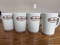 Front view of four white ceramic Tim Horton coffee mugs in a row on a wooden surface showing each mug's front Tim Horton logo in brown oval.