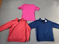 Three garments laid flat on carpet: pink short sleeve shirt, coral red 1/4 zip sweatshirt, navy blue 1/4 zip sweatshirt.