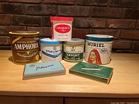 Full view of 5 tobacco containers and 2 cigarette packs on table against brick wall background