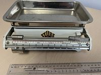 Front view of the Stube kitchen scale showing the brand name and the weighing tray on top. The sliding scale marks are clearly visible with the weight range in kilograms and grams.