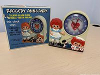 Front view of vintage Raggedy Ann and Andy talking alarm clock next to original blue box with yellow and white lettering and an image of the clock on the front.