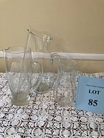 Set of three clear glass pitchers with textured design on lace tablecloth, includes stirring rod