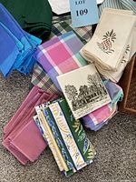 Photo of assorted textile napkins and tablecloths in blue, pink, green, plaid, floral and scenic prints folded together with a woven basket partially visible.
