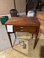 Front view of closed Singer Model 88 sewing table showing flat wooden top, two small drawers, tapered legs with gold-tone accents, foot pedal and power cord on floor.
