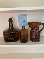 Wide shot of three brown bottles including Great Western wine bottle, E. G. Booz's Old Cabin Whiskey replica bottle, and brown Majolica pitcher.