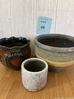 Three vintage-style flower pots of different sizes displayed on wooden surface against white wood paneled background. From left to right: dark brown glossy pot with embossed floral design, medium textured pot with band pattern, small white pot with crackle glaze.