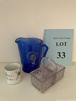 Overview of all three items including the souvenir cup, cobalt blue Shirley Temple pitcher, and clear glass basket with a 'LOT 33' card behind.