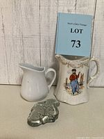 Photo showing all three items: small white ceramic pitcher, larger ornate ceramic pitcher with Dutch sailor print, clear glass fleur-de-lis paperweight.