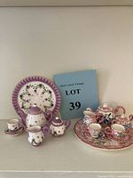 Photo showing both miniature child's tea sets side by side, displaying teapots, cups, saucers, sugar bowl, creamer, and tray.