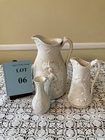 Photo of all three ceramic items: large Wedgwood Jasperware pitcher, medium US Pottery Co Parian pitcher, and small white parian vase, arranged on crocheted lace.
