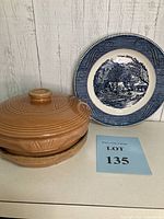 Angle showing front view of brown pottery casserole dish with matching lid and Currier & Ives decorative plate.