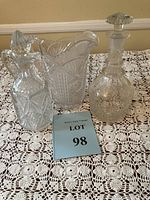Photo of the three clear-cut vintage glass vessels displayed on a crocheted lace tablecloth including decanter with stopper, pitcher or vase, and carafe/bottle