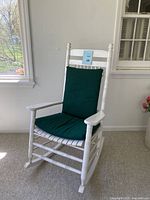 White wooden rocking chair with slat seat and green cushion positioned near window and white wall