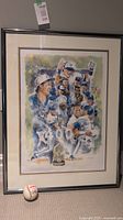 Framed and matted artwork showing group of Toronto Blue Jays players celebrating with championship trophy at bottom center, with baseball placed on floor beside frame.