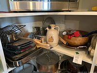Photo of a kitchen shelf containing stacked glass baking dishes, wooden cutting boards, ceramic oil pitcher, wooden salad bowl with utensils, metal strainer and several metal pots and pans.