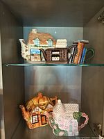 Five ceramic teapots shaped like houses, displayed on two shelves inside a cabinet, showing detailed house designs and colors
