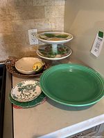 Wide view of various ceramic serving dishes on counter including two cake stands, lemon dish, brown dish, small dishes, and large green Fiesta plate