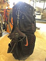 Front view of black Nike golf stand bag with orange and white swoosh logo, zippered storage pockets, and built-in stand.