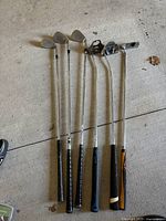 Six left-handed golf clubs including irons and putter laid on concrete floor, showing black grips and steel shafts