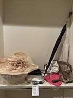 Photo of beige wide-brim ladies fancy hat with floral decoration, multiple small handbags, black umbrella, and vanity dresser set items on shelf