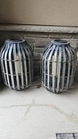 Pair of metal strapped patio lanterns placed on concrete floor against stone wall corner, showing weathered surfaces and vertical metal straps with wooden bases and tops.