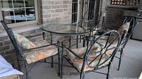 Patio dinette set showing round glass top table with four cast metal chairs with floral cushions on outdoor concrete floor by brick wall.