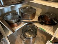 Upper shelf shows stainless steel Lagostina stock pot with glass lid; lower shelf shows stainless steel pot with steamer insert and partial view of copper cookware.